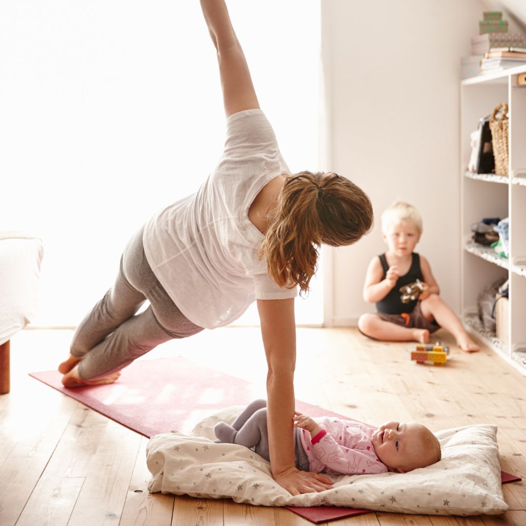 young mom practicing yoga with infant on yoga mat and toddler playing in the background.